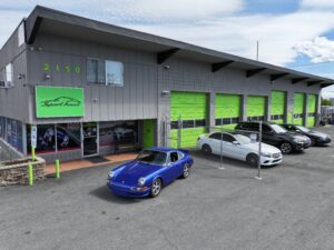 Exterior view of Sport Haus Reno European auto repair shop at 2150 Mill St, Reno, NV 89502, featuring a lineup of luxury vehicles including a vintage blue Porsche and multiple Mercedes-Benz cars, with bright green service bay doors and clear signage.