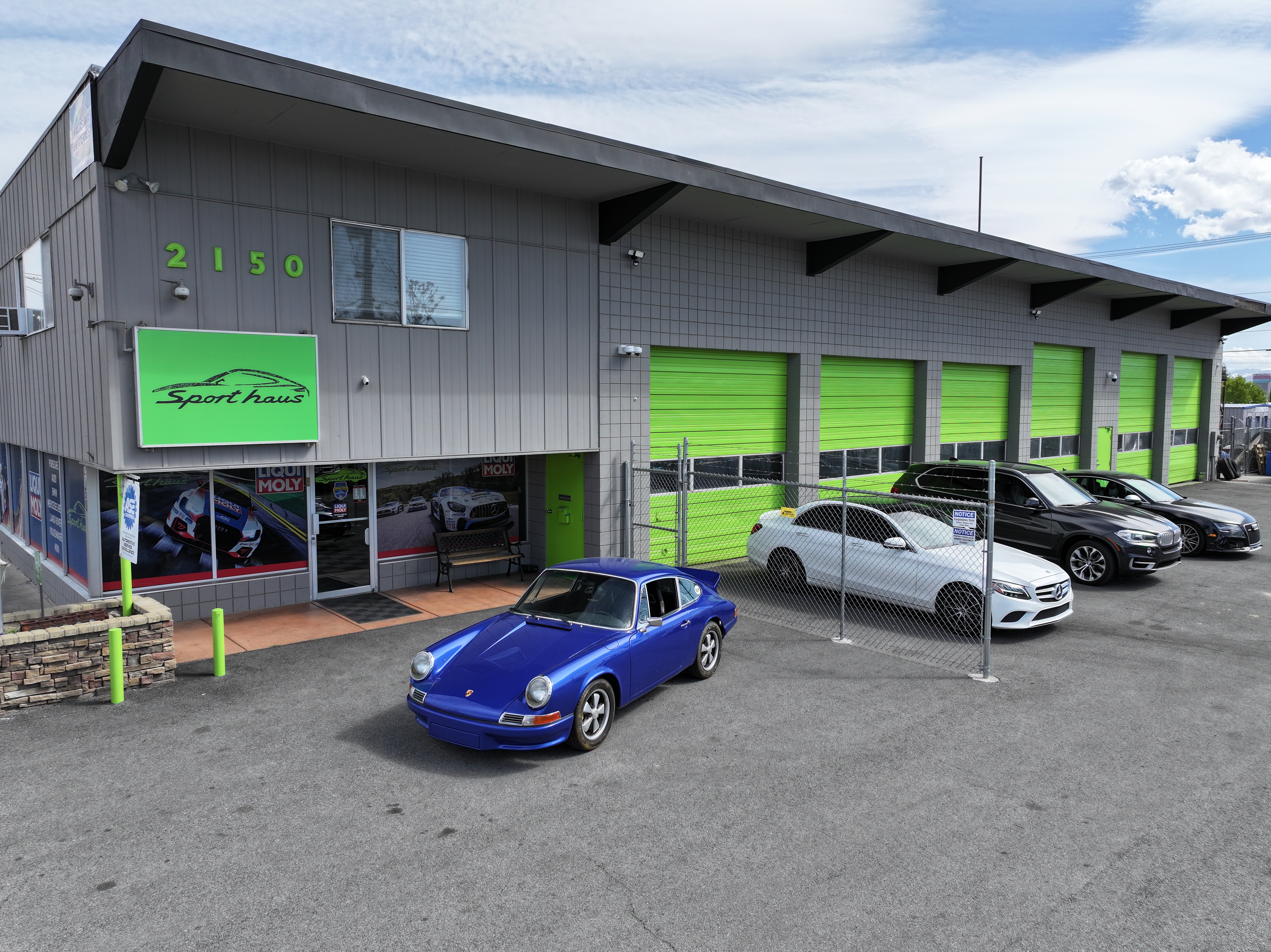 Exterior view of Sport Haus Reno a European car mechanic near me auto repair shop at 2150 Mill St, Reno, NV 89502, featuring a lineup of luxury vehicles including a vintage blue Porsche and multiple Mercedes-Benz cars, with bright green service bay doors and clear signage.